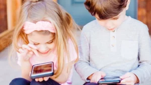 Two young children are engrossed in smartphones, with the girl smiling as she interacts with her device, and the boy focused intently on his screen. This image reflects the considerations parents must weigh regarding smartphone readiness for kids, a topic discussed in the blog "Is Your Child Ready for a Smartphone?"