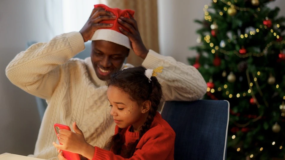 A joyful father watches his daughter engrossed in her new device on Christmas, highlighting the importance of discussing device usage and safety
