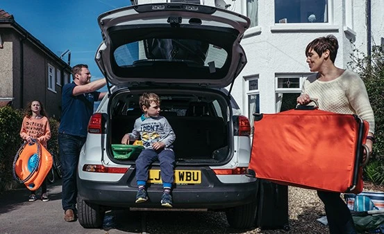 A family is busy loading their car for a road trip, with a young boy sitting in the open trunk holding a toy, a woman carrying a large orange bag, a man arranging items, and a girl holding a floatie, symbolizing preparations for a long journey filled with activities and possibly listening to the '10 Best Kids Podcasts for Long Summer Road Trips'.