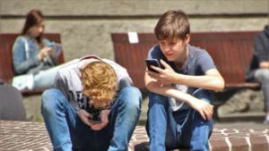 Two teenage boys are sitting outdoors, each engrossed in their own smartphones. The boy on the left, with ginger hair, is hunched over his phone, which he holds in his hands between his knees, possibly indicating intense focus or concern. The boy on the right, wearing a grey T-shirt and blue jeans, is sitting more relaxed with one arm resting on his bent knee, looking at his phone screen. In the background, a girl is sitting on a bench, also using her phone, illustrating the common scene of young people absorbed in their digital devices. The setting appears to be a public outdoor area, possibly a park or a school courtyard.