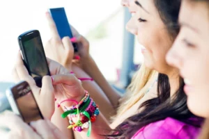 Three young individuals are intently looking at their smartphones, seemingly engaged in digital interaction. They are closely seated together, each holding a device. The focus on their screens suggests a connection to the broader digital community, potentially highlighting the modern youth's inclination towards constant connectivity and social media engagement. Visible accessories like colorful bracelets add a casual and youthful vibe to the scene.