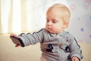 A curious toddler is holding a smartphone, symbolizing the early introduction to technology in today's digital age and the importance of parental guidance in online communications.